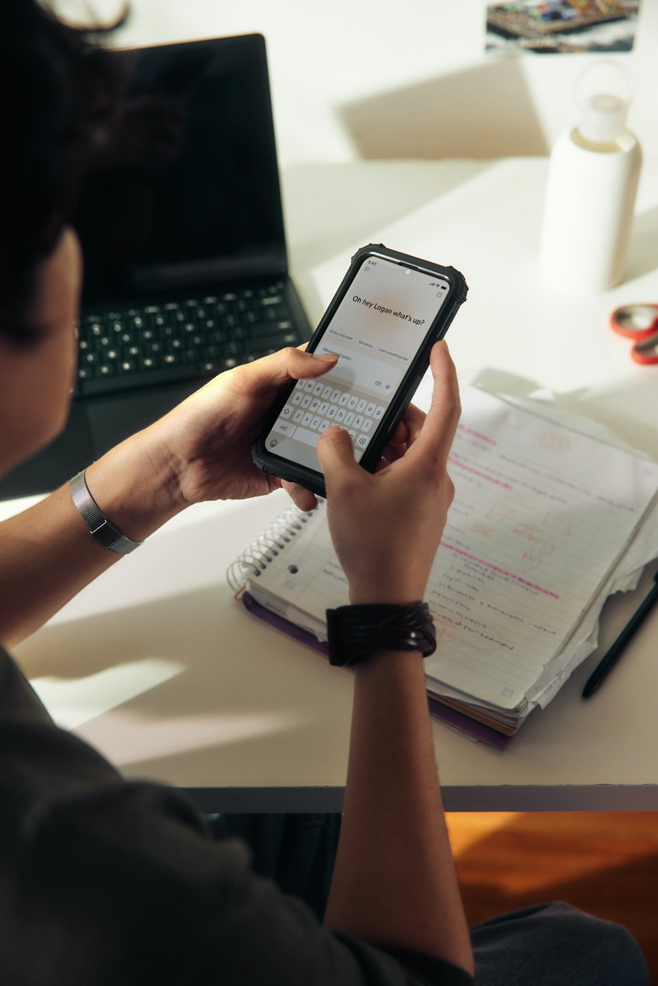 Hände tippen auf Smartphone neben Laptop und Notizbuch auf Schreibtisch.